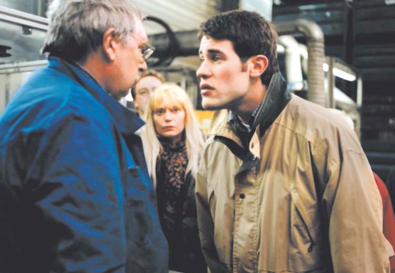 Still des Films RESSOURCES HUMAINES (F/UK 1999): In einer Fabrikhalle stehen zwei männlich gelesene Personen einander konfrontativ gegenüber. Im Hintergrund sehen mehrere Personen zu, darunter eine weiblich gelesene Person in der Bildmitte.